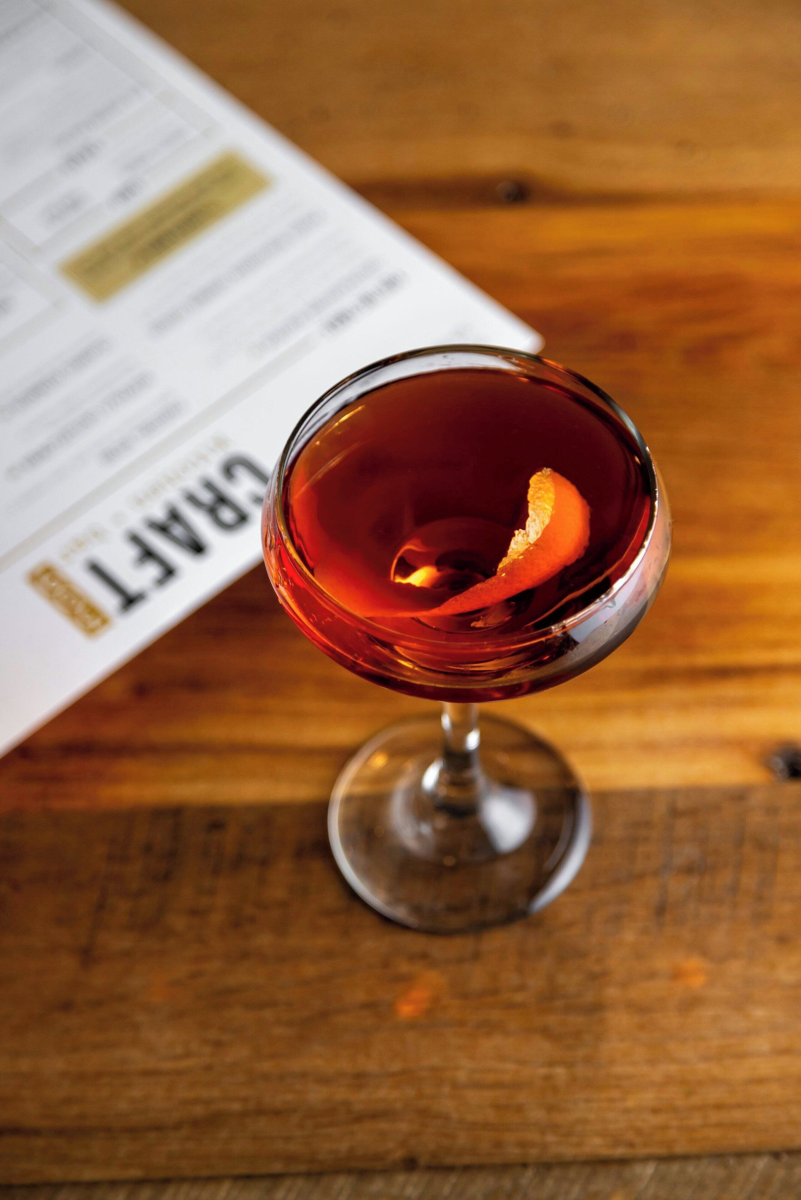 Een glas rode drank met een sinaasappelschil als garnering, op een houten ondergrond, met een gedeeltelijk zichtbaar menu op de achtergrond.