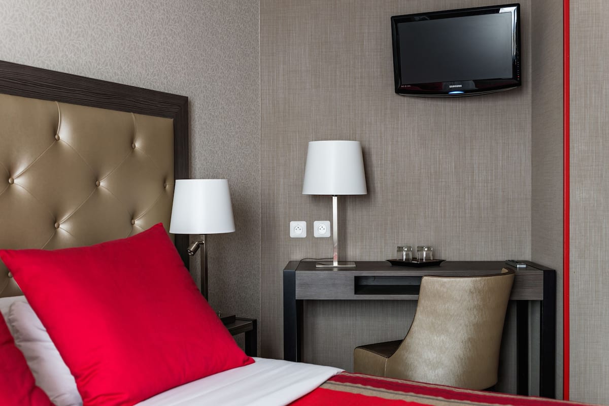 A hotel room with a tufted headboard, a bed with red and white bedding, a desk with a lamp, a chair, and a flat-screen TV mounted on the wall.