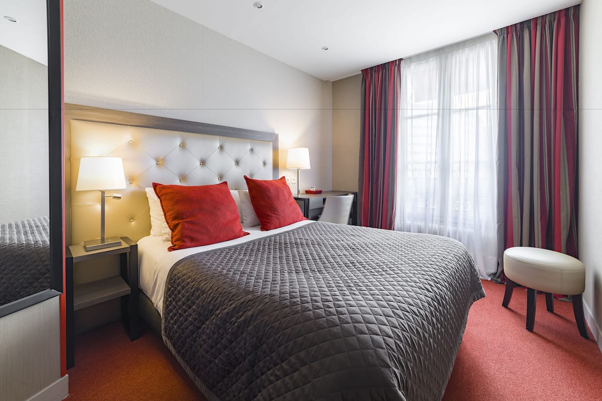 A hotel room with a large bed, two bedside tables with lamps, a red accent pillow, a white bench, and a window with curtains.