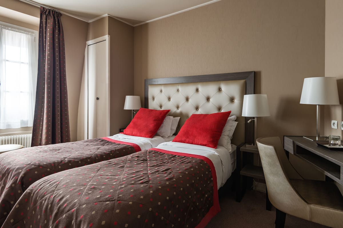 A hotel room with two beds, each with red and white bedding, a large tufted headboard, bedside lamps, and a desk with a chair.