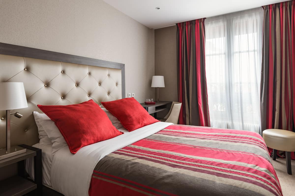 A hotel room with a tufted headboard, a bed with red and white bedding, a nightstand with a lamp, a desk with a lamp, and a window with striped curtains.