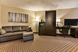 A hotel room with a patterned carpet, a sofa, a desk with a computer, a television, and a cabinet.