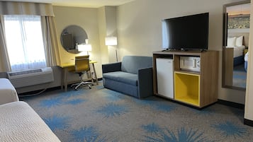 Et hotelværelse med seng, skrivebord med stol, sofa, fjernsyn, lille køleskab og et spejl.