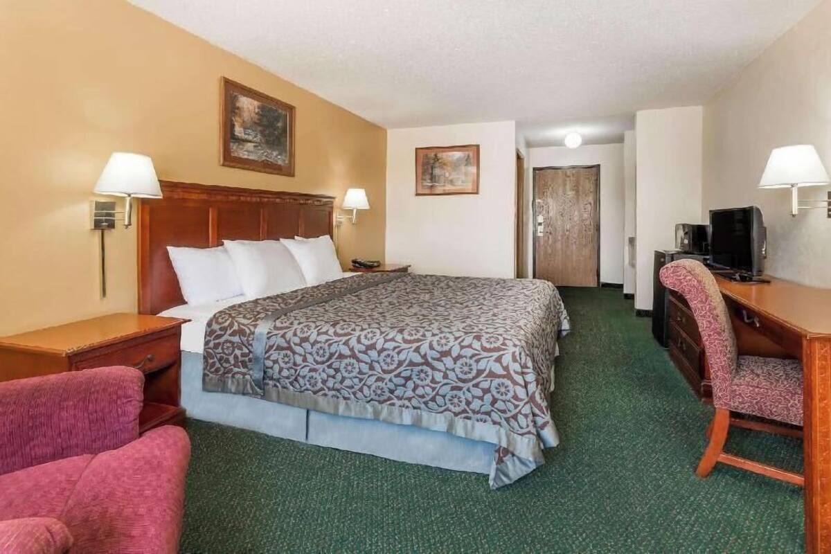 A hotel room with a bed, a desk, a chair, a lamp, and a door.