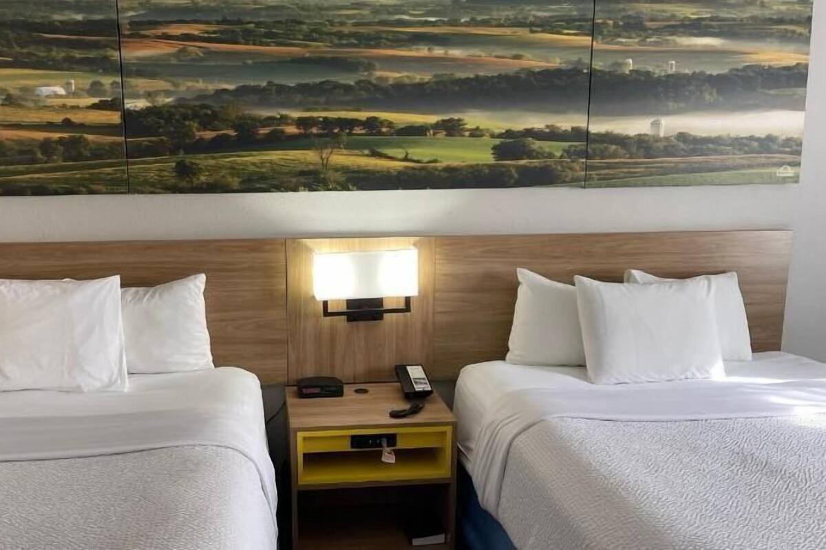 A hotel room with two beds, a nightstand with a lamp, and a large landscape painting above the beds.