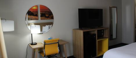 A hotel room with a bed, a desk with a chair, a television, a mirror, and a wall-mounted artwork.