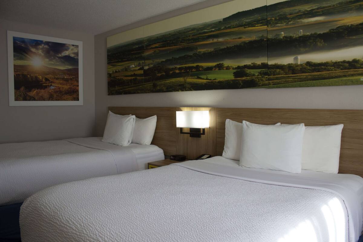 A hotel room with two beds, a large landscape painting above the beds, and a nightstand with a lamp.