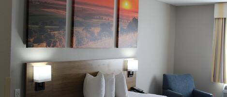 Habitación de hotel con cama, lámparas de noche, mesita de noche, una silla y tres cuadros en la pared.