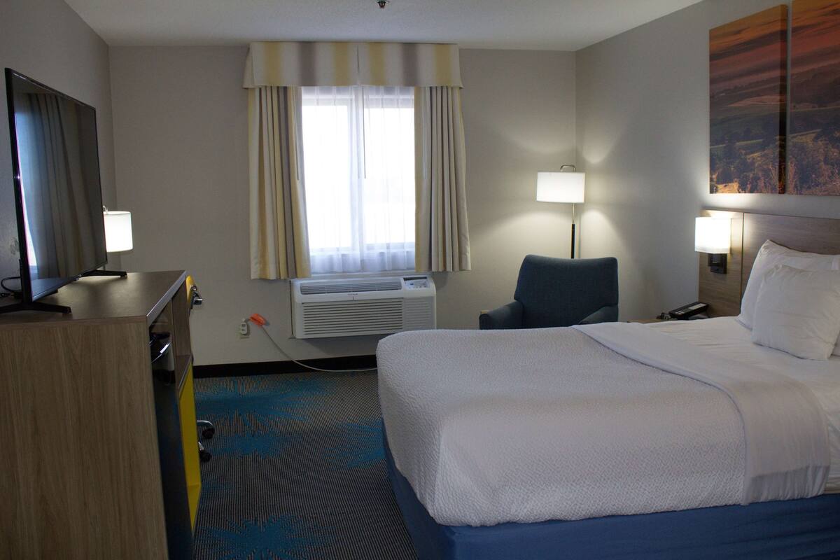 A hotel room with a bed, a desk, a chair, a TV, a window with curtains, an air conditioner, and a lamp.