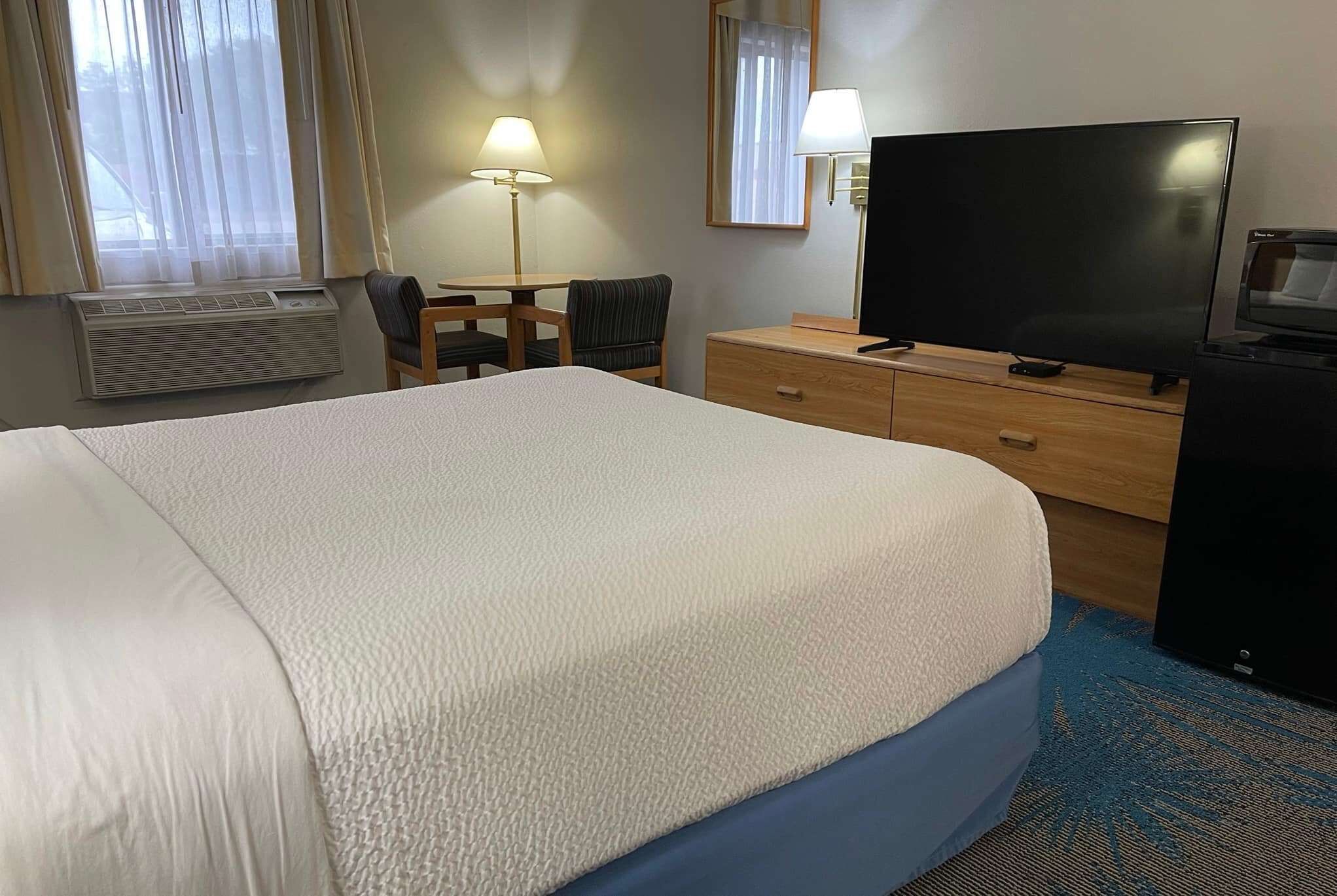 A hotel room with a large bed, a desk with a chair, a television, a dresser, and an air conditioning unit.
