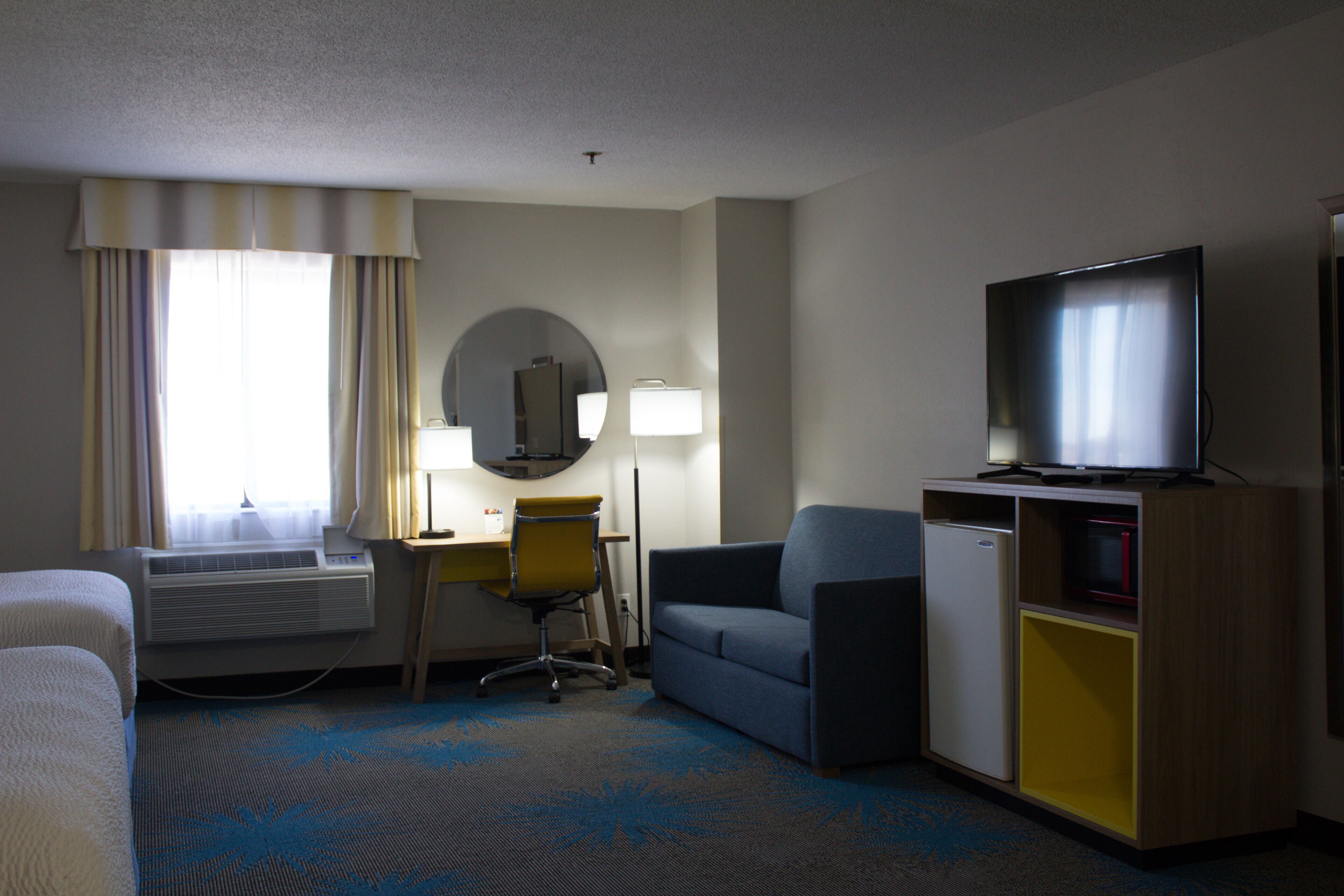 A hotel room with a bed, a desk with a chair, a television, a small refrigerator, and a mirror.