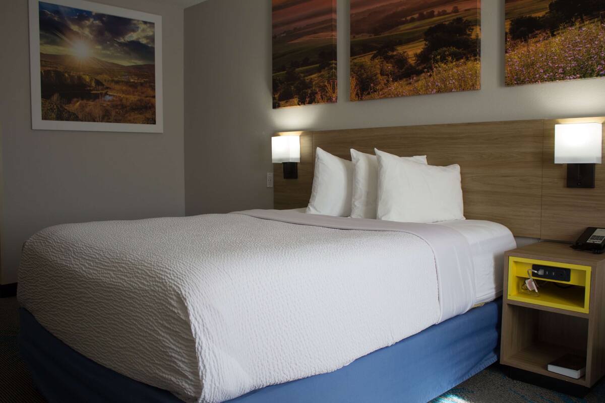 A hotel room with a large bed, two bedside lamps, a wooden headboard, and three landscape paintings on the wall.