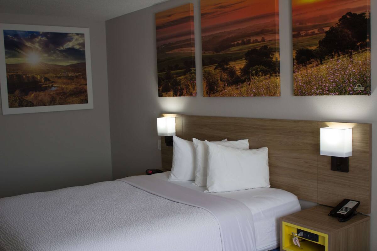 A hotel room with a bed, two wall-mounted lamps, a bedside table with a phone, and three landscape paintings on the wall.
