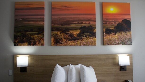 A hotel room with a bed, two bedside lamps, a nightstand with a phone, and three sunset paintings on the wall. - Days Inn by Wyndham Colby (Colby)