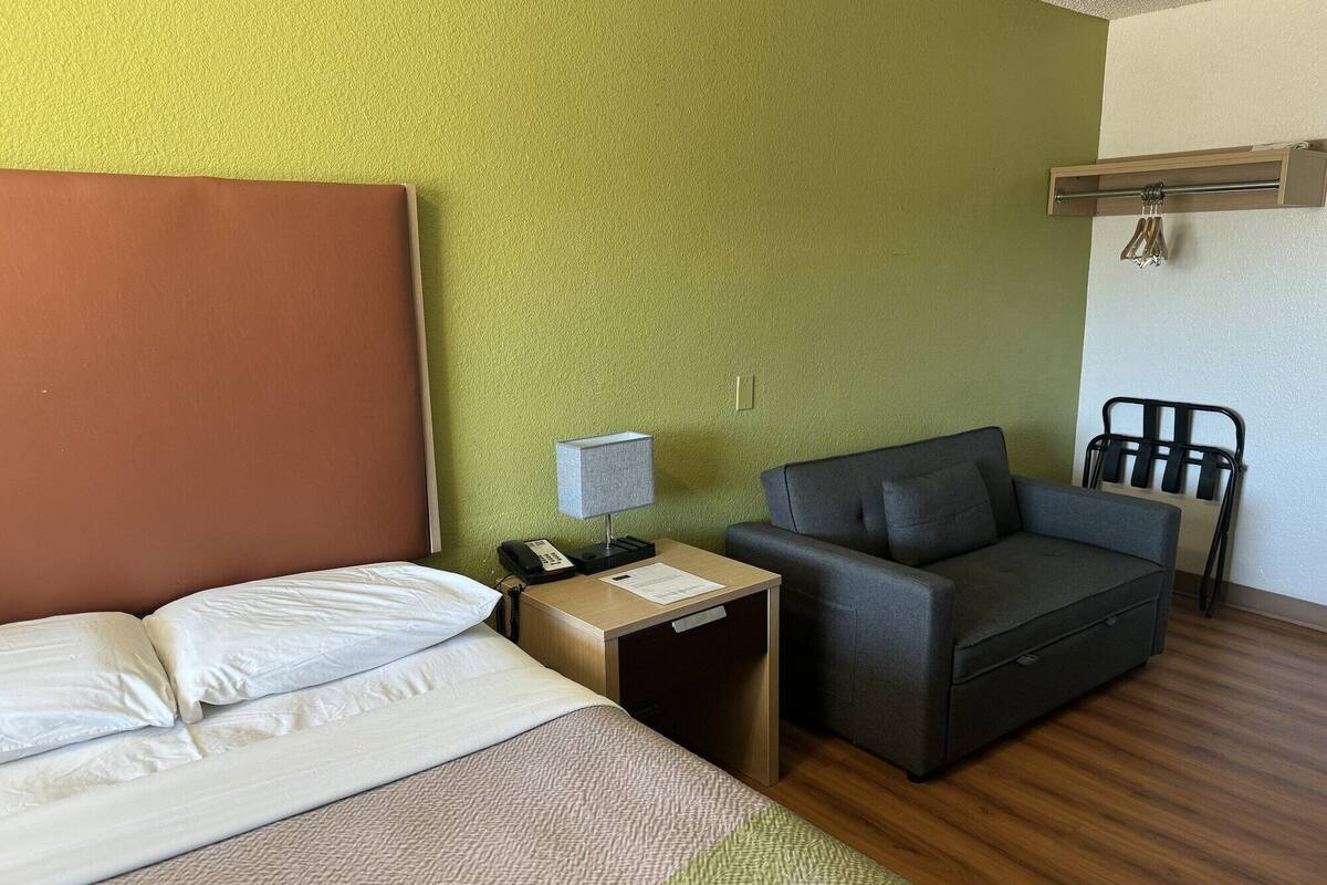 Ein Hotelzimmer mit einem Bett, einem Schreibtisch mit Lampe, einem grauen Sofa, einem Sessel und einem hölzernen Kopfteil.