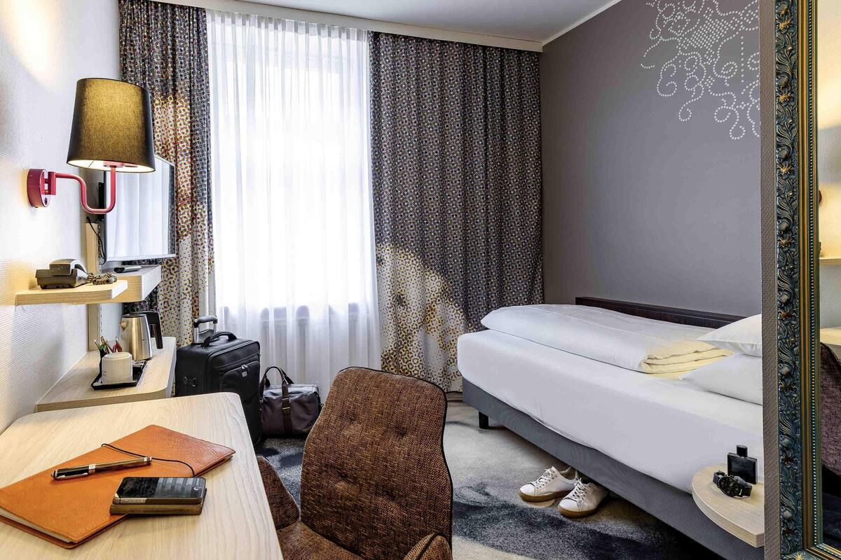 Quarto de hotel com cama, escrivaninha, cadeira, abajur e espelho.