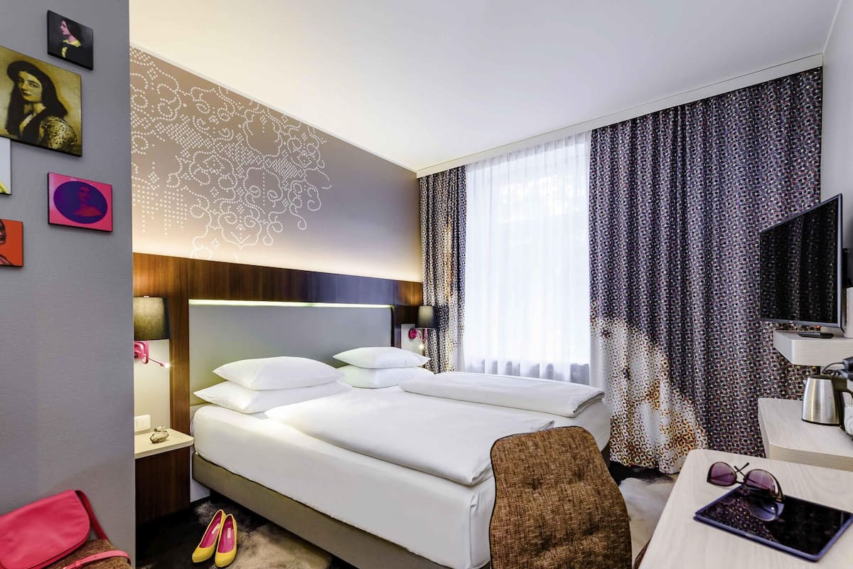 Quarto de hotel com cama, mesa de cabeceira, televisão e escrivaninha com cadeira.