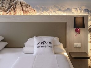A hotel room with a bed, a wall-mounted lamp, and a view of mountains. - Mercure Hotel Muenchen am Olympiapark (Munich)