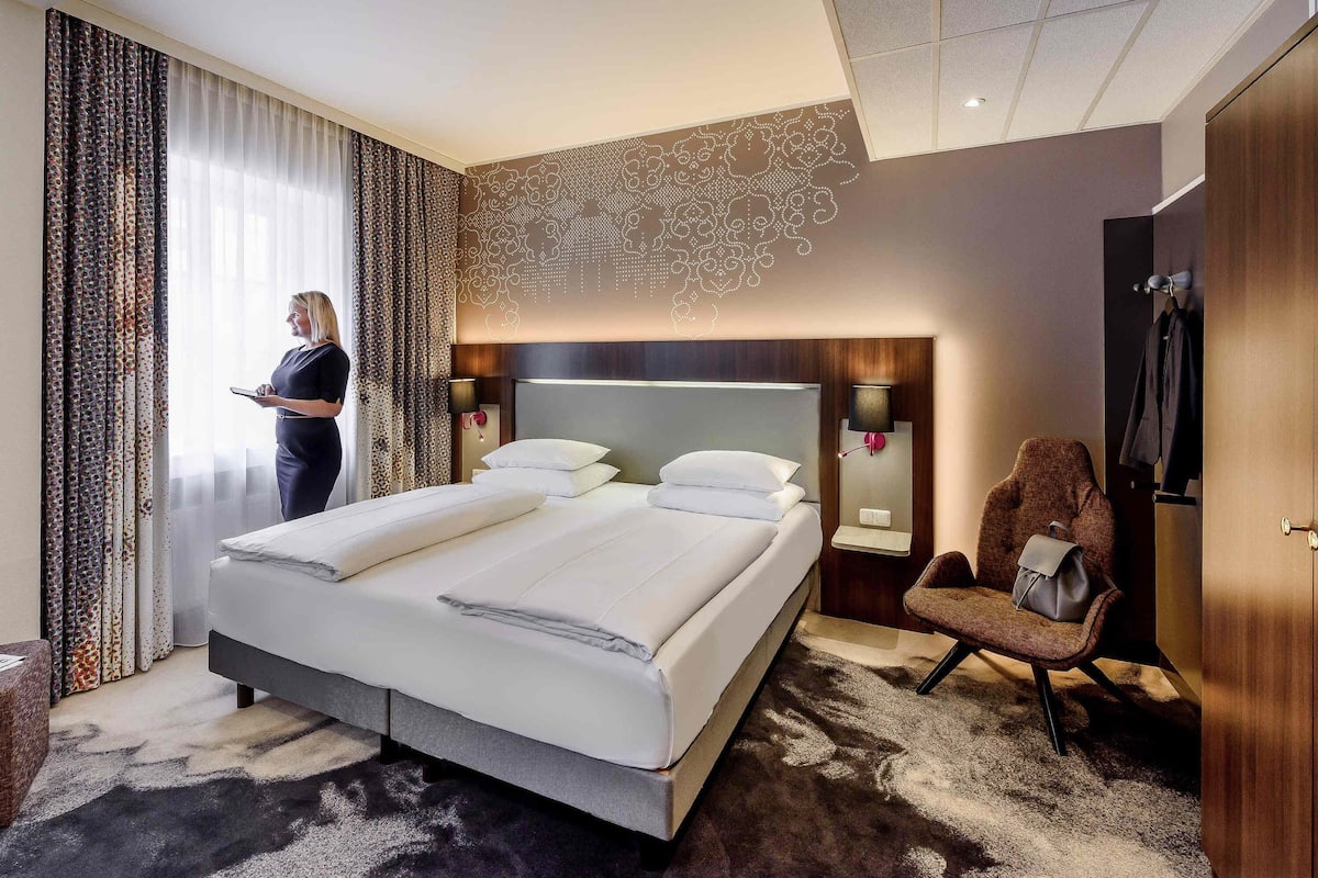Quarto de hotel com uma cama grande, luminárias de cabeceira, uma cadeira e um cabideiro.
