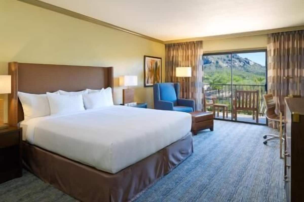 Habitación de hotel con una cama grande, un escritorio, una silla y un balcón con vistas a la montaña.