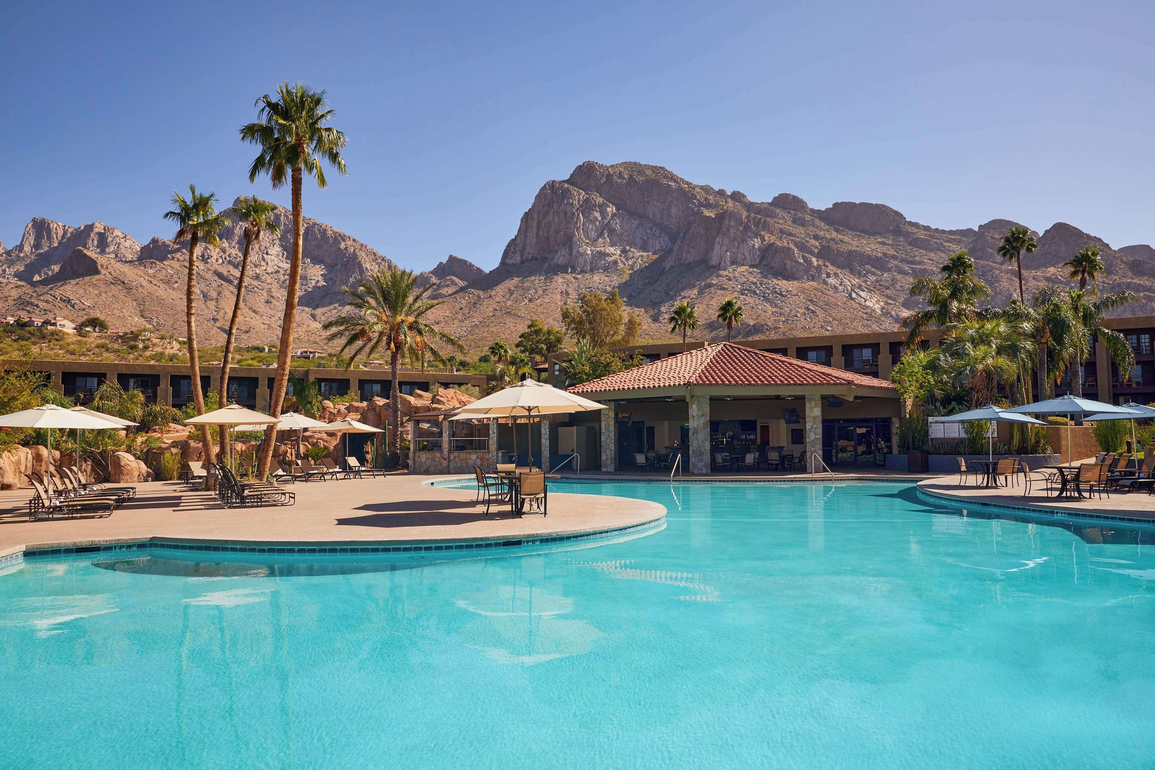 Une piscine de complexe hôtelier bordée de palmiers, avec une chaîne de montagnes en arrière-plan.