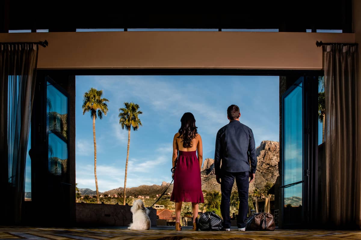 Una pareja de pie frente a un umbral abierto, con vista a un paisaje con palmeras y montañas.