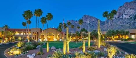 A hotel with a desert garden featuring cacti and palm trees, a mountain backdrop, and a well-lit entrance.