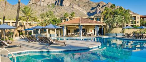 A resort pool with lounge chairs, umbrellas, and a mountain backdrop.