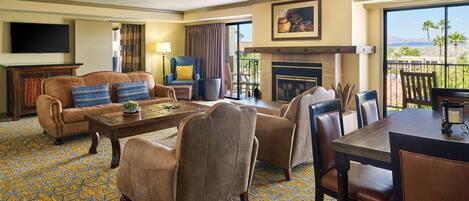 Un salón de hotel con sofá, sillones, una mesa de centro, chimenea, televisor y un balcón con vistas a palmeras y montañas.