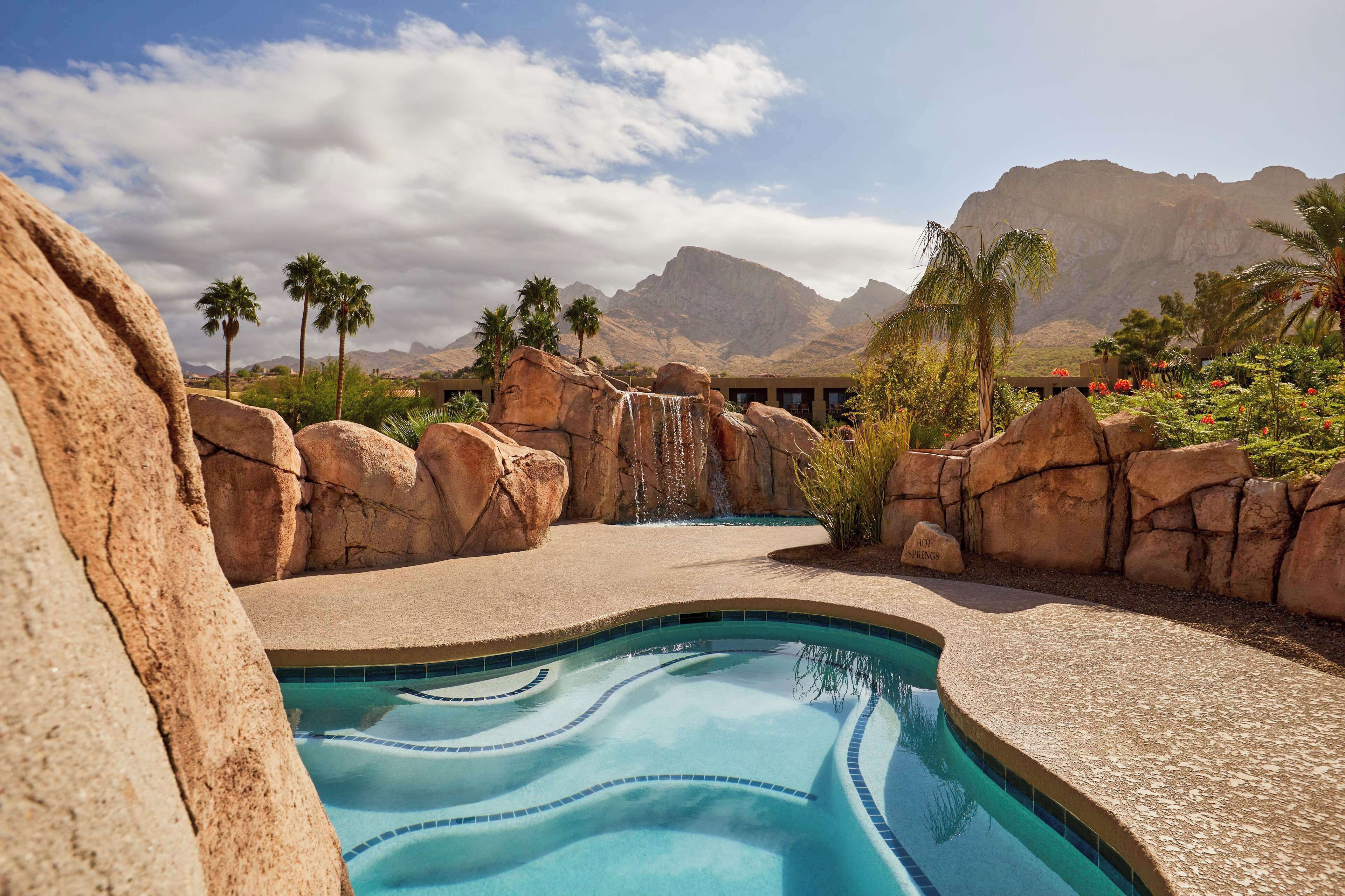 A pool area with a waterfall, surrounded by large rocks and palm trees, with mountains in the background.