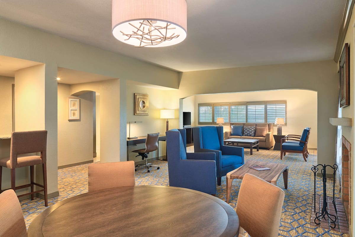 Habitación de hotel con una mesa de comedor redonda, sillones azules, un sofá, un escritorio con silla y un ventanal con persianas.