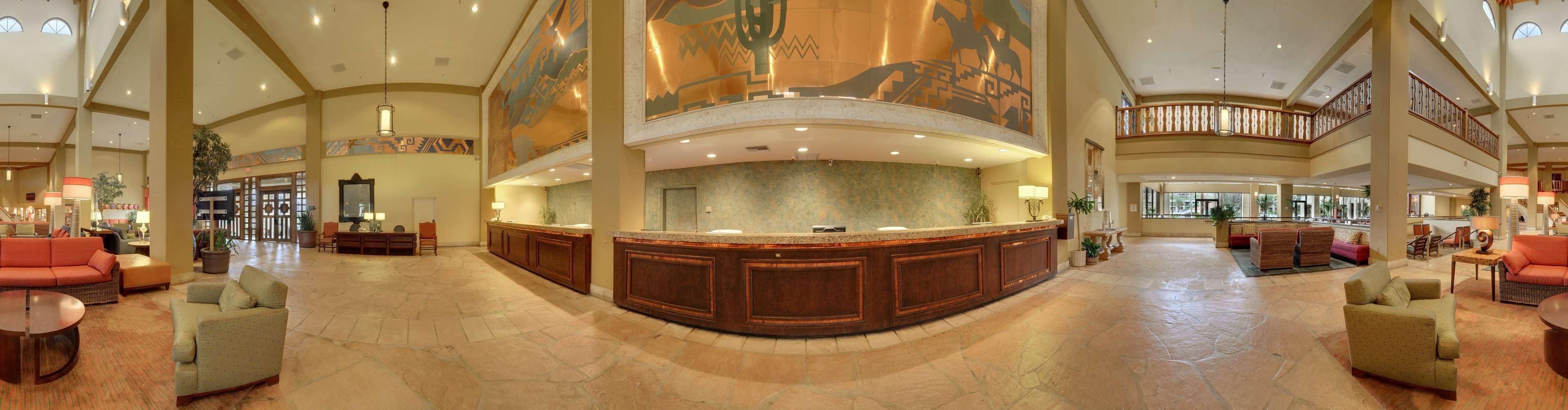 Um hall espaçoso com um balcão de recepção central, áreas de estar e um teto alto com murais decorativos.