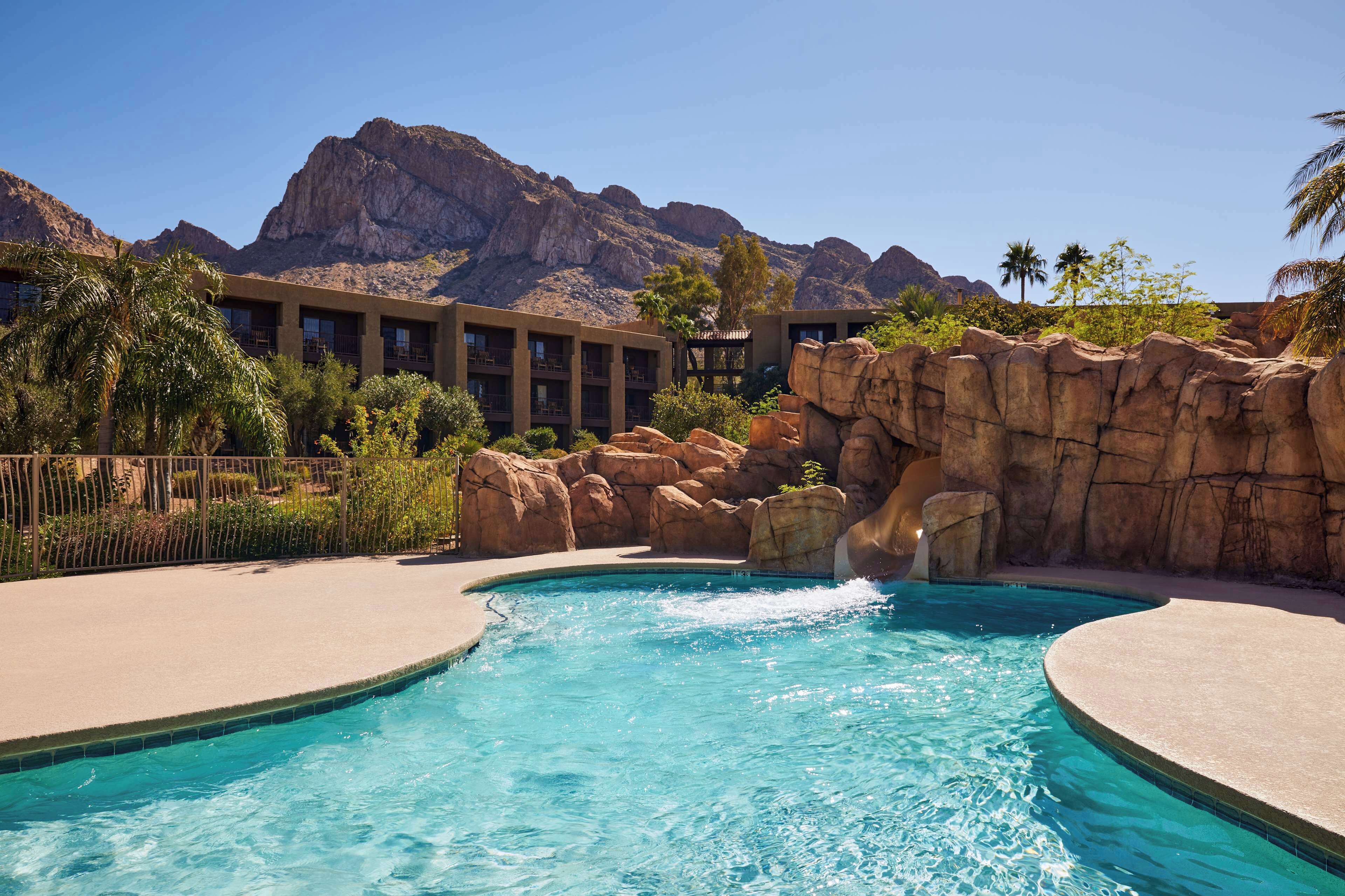 A pool with a waterfall feature, surrounded by rock formations and palm trees, with a mountain backdrop.