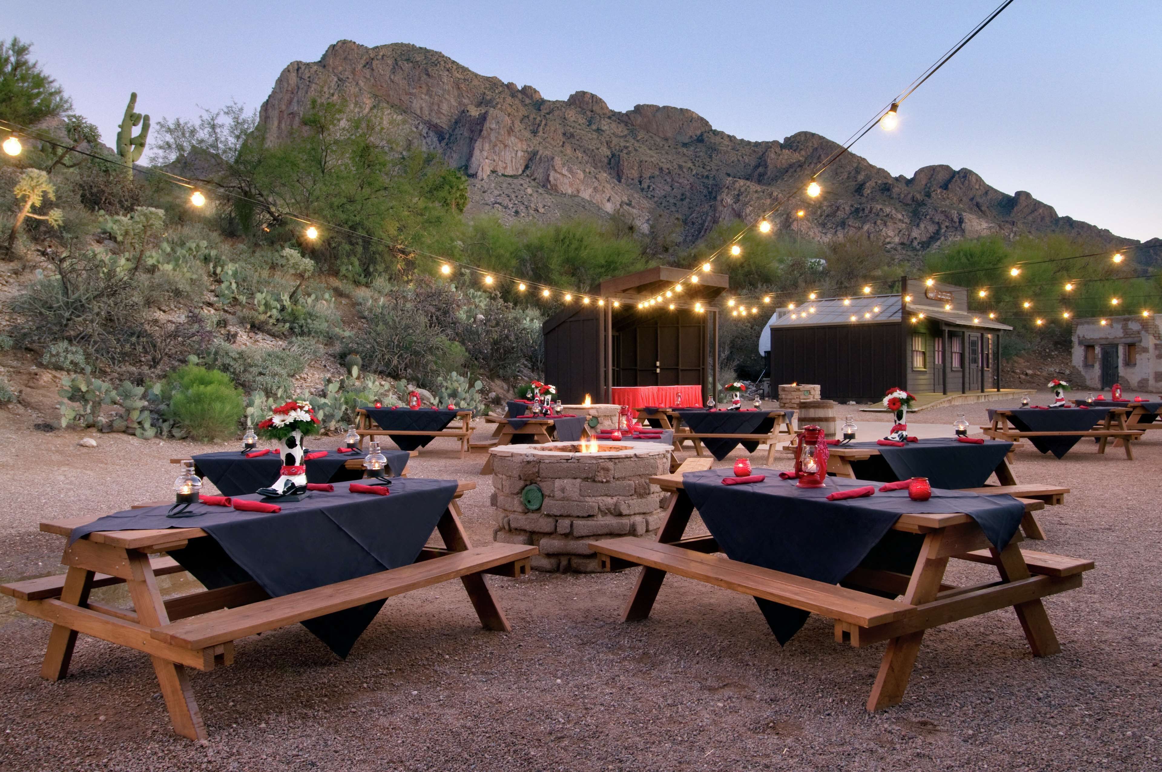 An outdoor dining area with wooden tables and benches, string lights, and a stone fire pit.