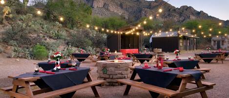 An outdoor dining area with wooden tables and benches, string lights, and a stone fire pit.