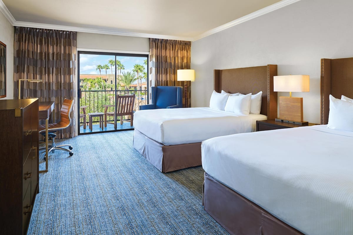 Habitación de hotel con dos camas, un escritorio, una silla y vistas a palmeras.