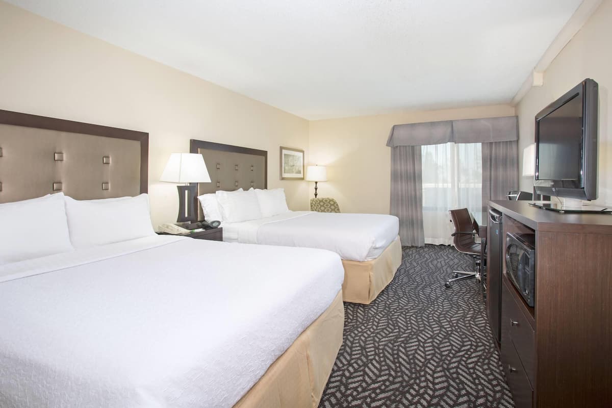 A hotel room with two beds, a desk, a television, and a large window with curtains.