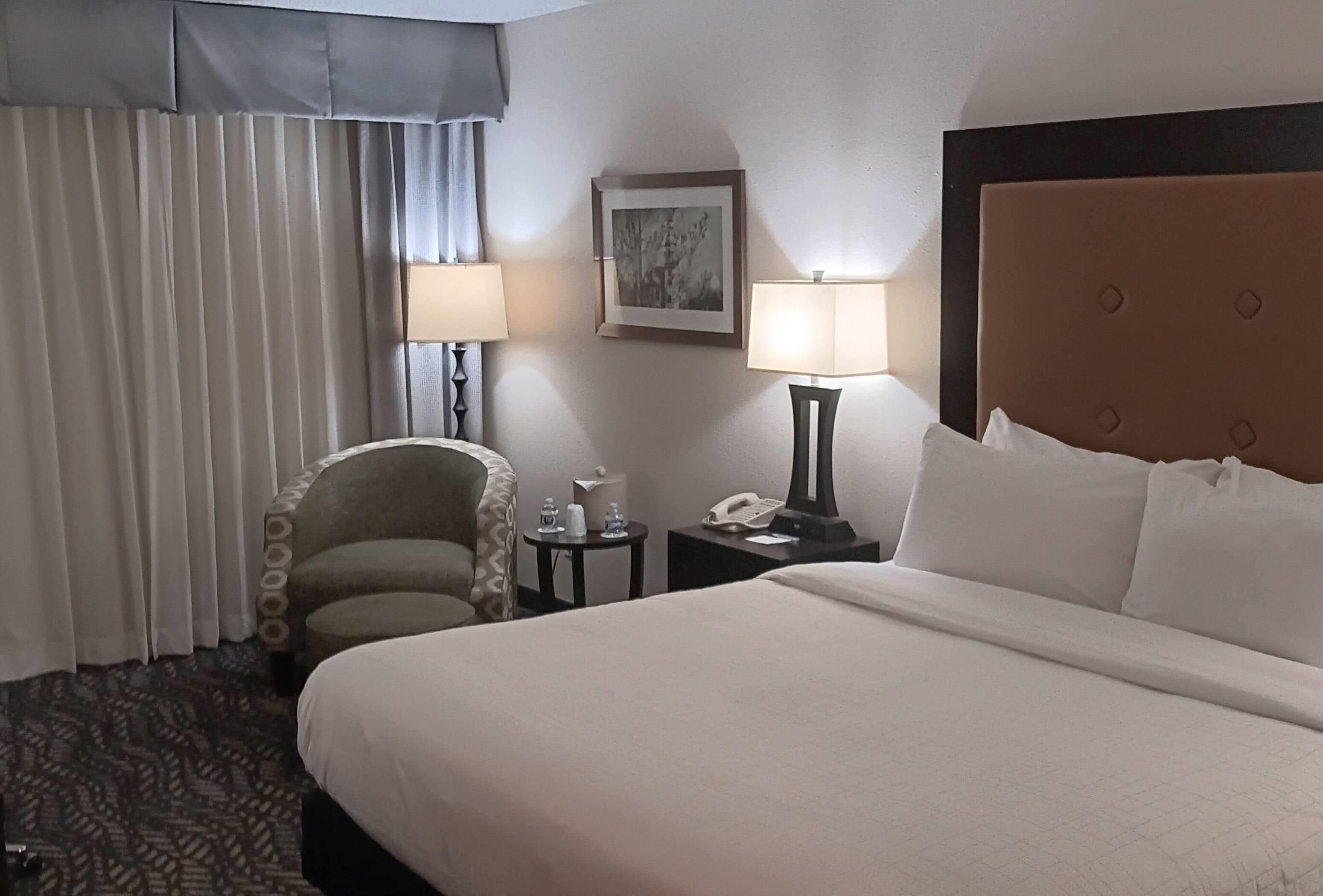 A hotel room with a bed, a chair, a nightstand with a lamp, a telephone, and a painting on the wall.