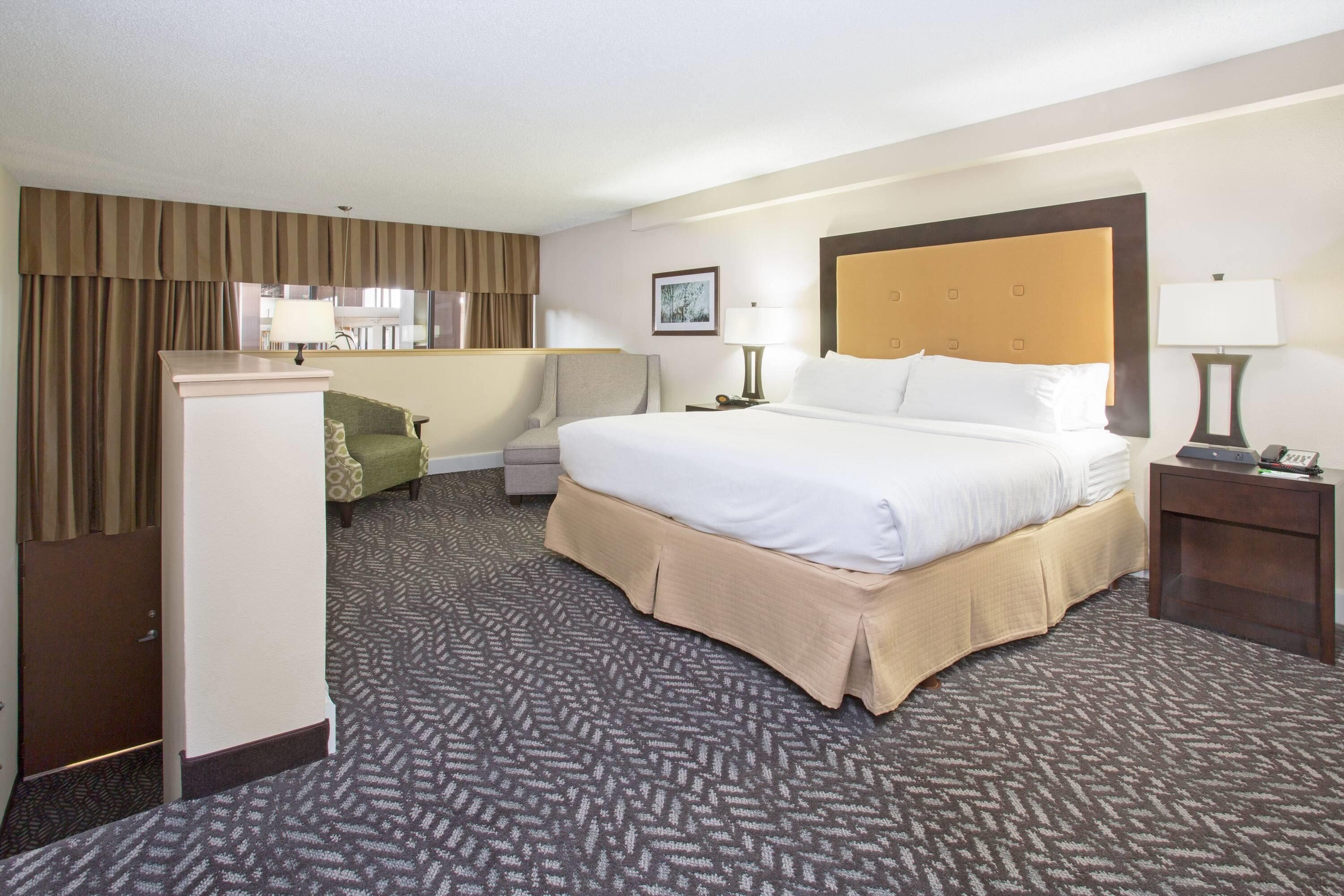 A hotel room with a large bed, a desk, a chair, a wardrobe, and a small table with a lamp.