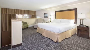 A hotel room with a large bed, a desk, a chair, a wardrobe, and a small table with a lamp.