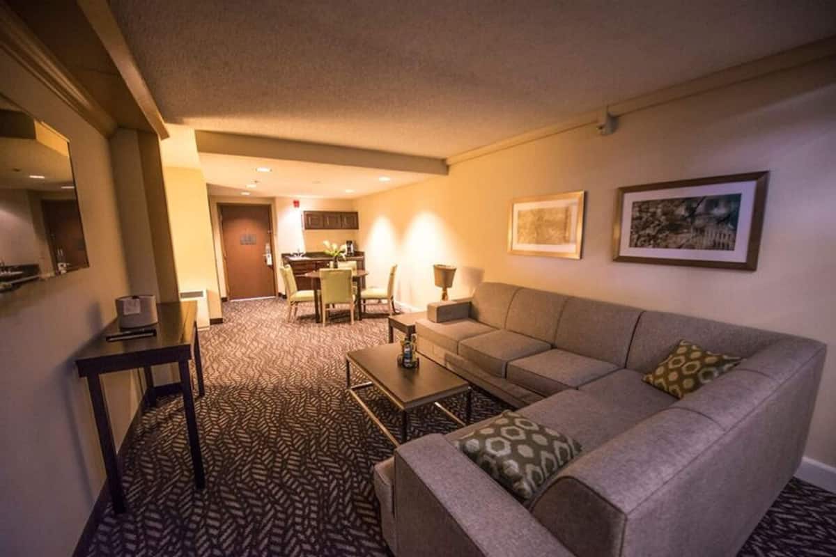 A hotel room with a sofa, a coffee table, a dining area with chairs, and a kitchen in the background.
