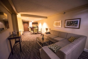 A hotel room with a sofa, a coffee table, a dining area with chairs, and a kitchen in the background.