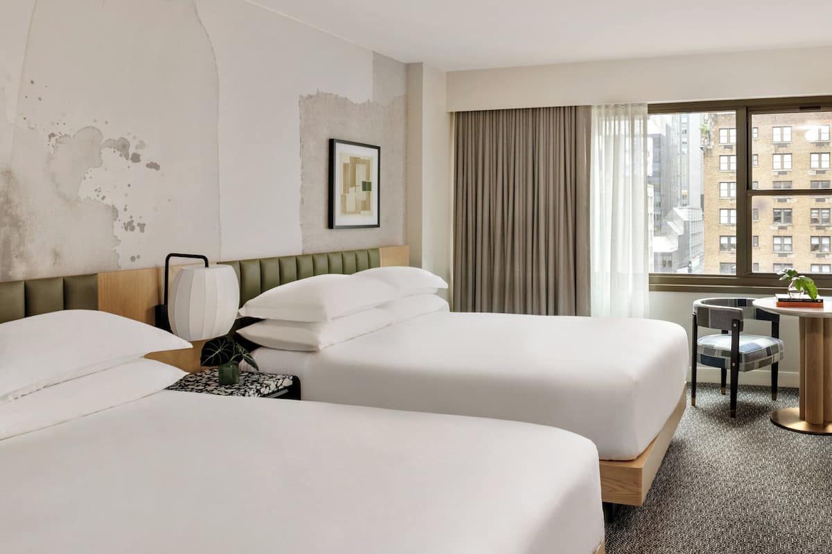 A hotel room with two beds, a small table with a plant, a chair, and a window with a view of buildings.