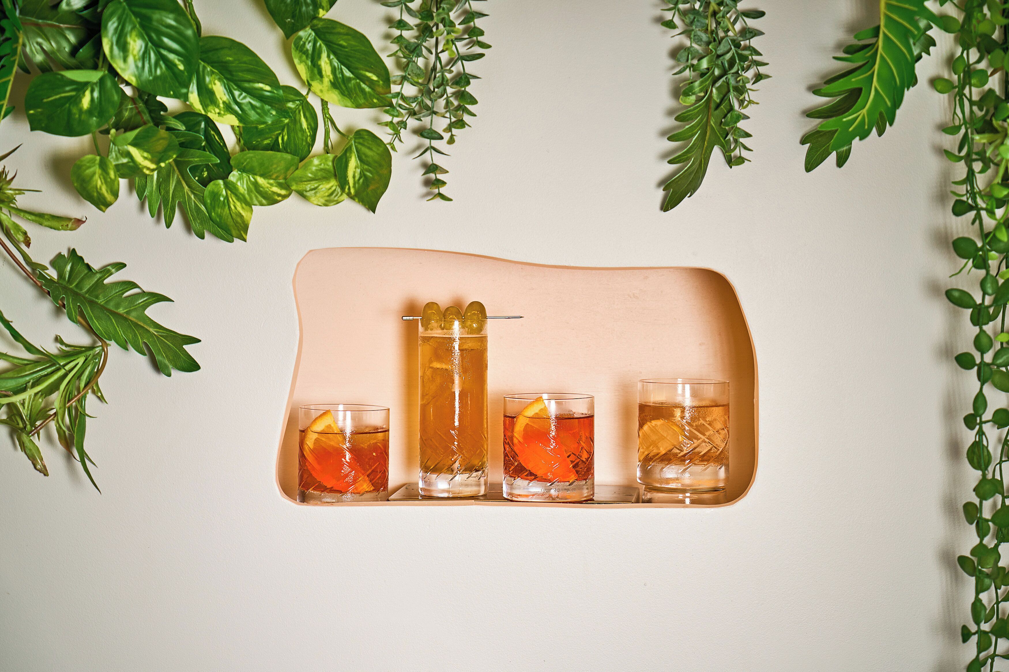 A tray with four glasses of amber-colored beverages, each with an orange slice, surrounded by green foliage.