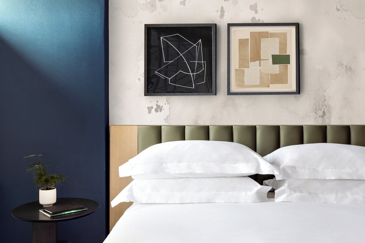 A neatly made bed with white linens, a round side table with a plant, and two abstract art pieces on the wall.