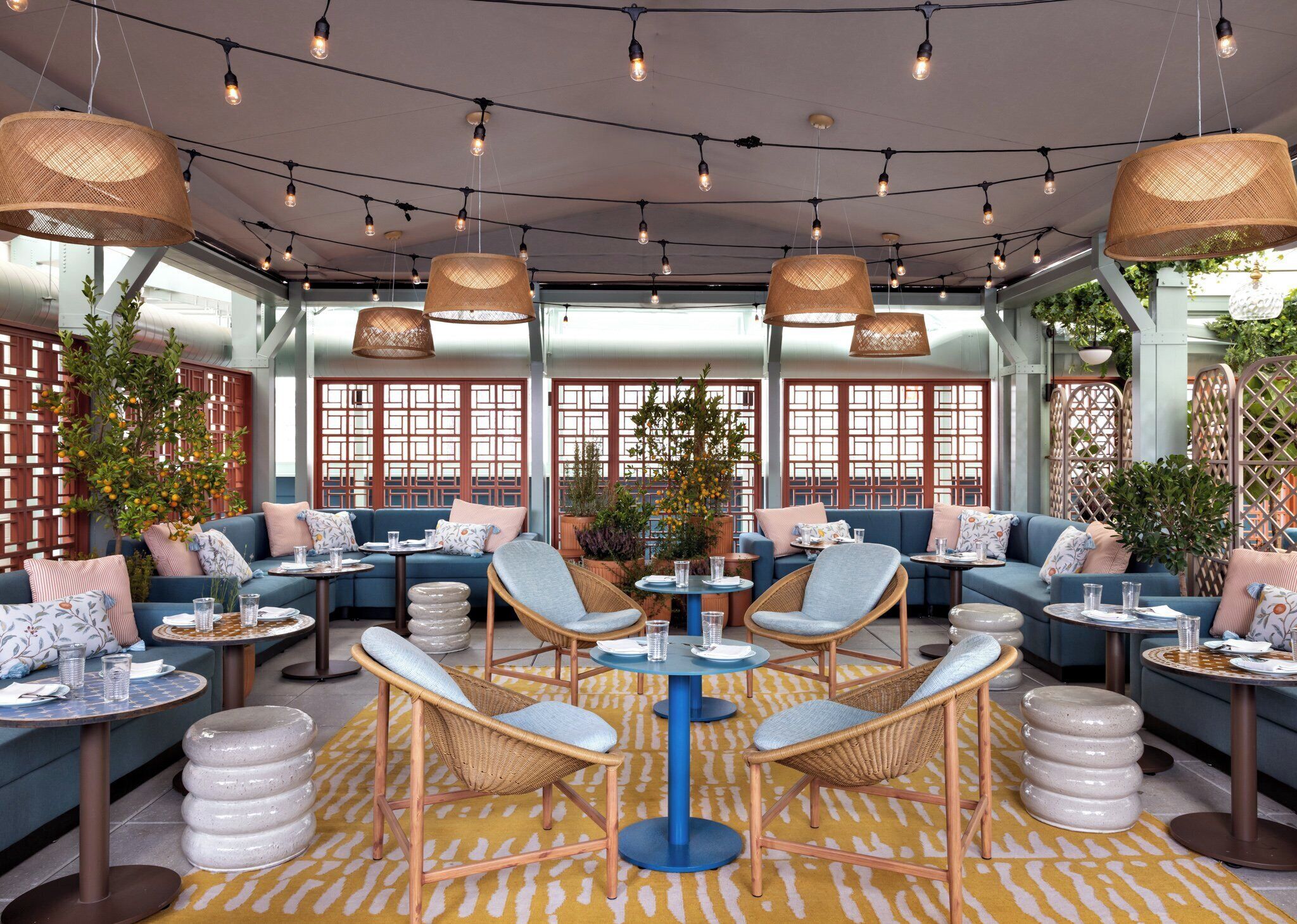 A modern lounge area with sofas, chairs, and tables, featuring a yellow patterned rug, wicker furniture, and string lights.