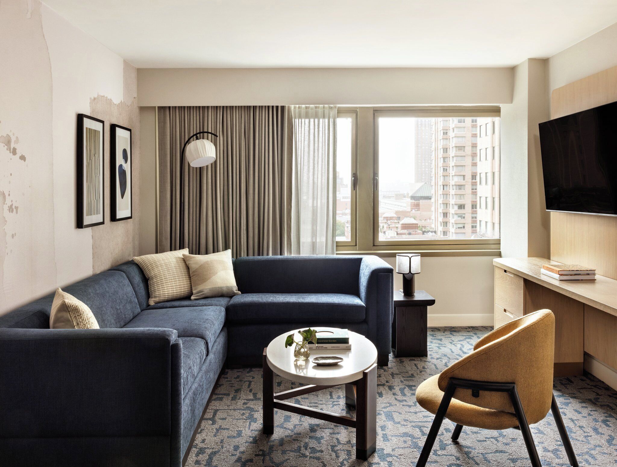 A modern hotel room with a sectional sofa, a round coffee table, a single armchair, a TV, and a large window with a city view.