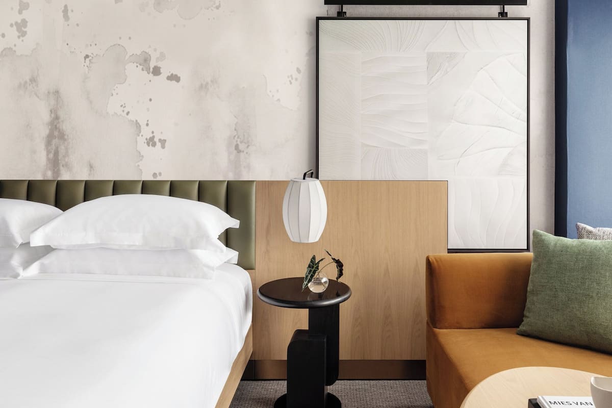 A hotel room with a bed, a sofa, a small table with a plant, a wall-mounted mirror, and a large abstract painting.