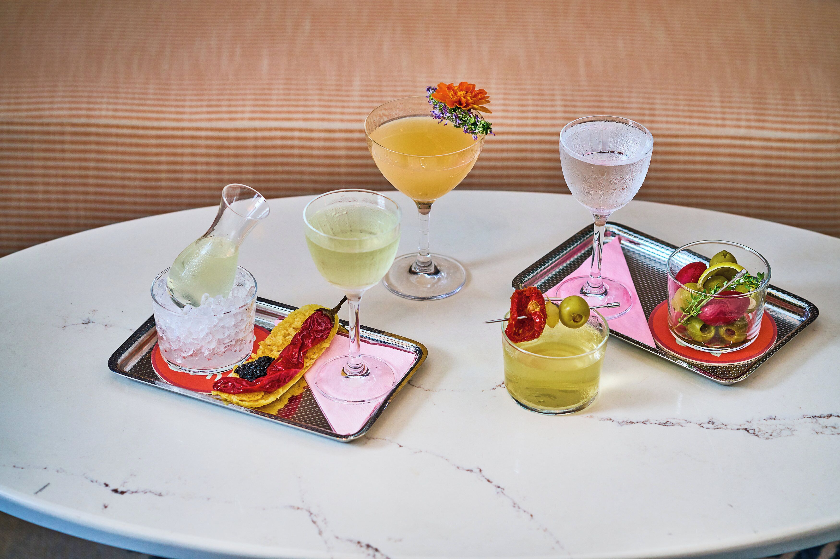 A table with various drinks and appetizers, including a cocktail with a flower garnish, a glass with a red garnish, and a glass with olives and a lemon.
