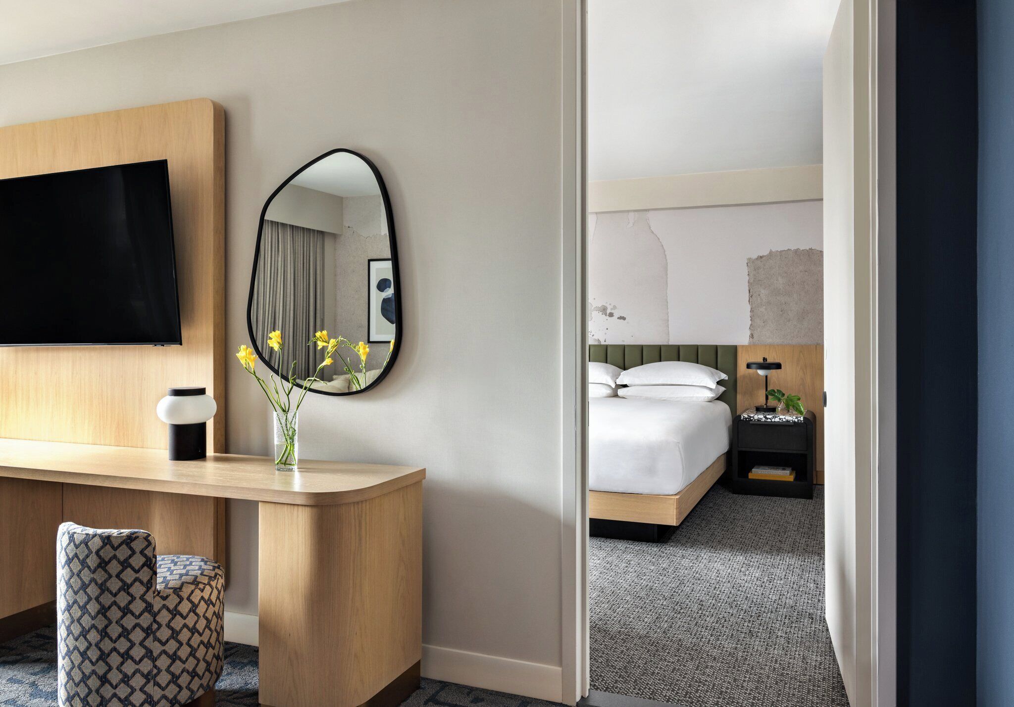 A hotel room with a wooden desk, a flat-screen TV, a round mirror, a vase with flowers, a bed with white bedding, and a nightstand with a lamp.